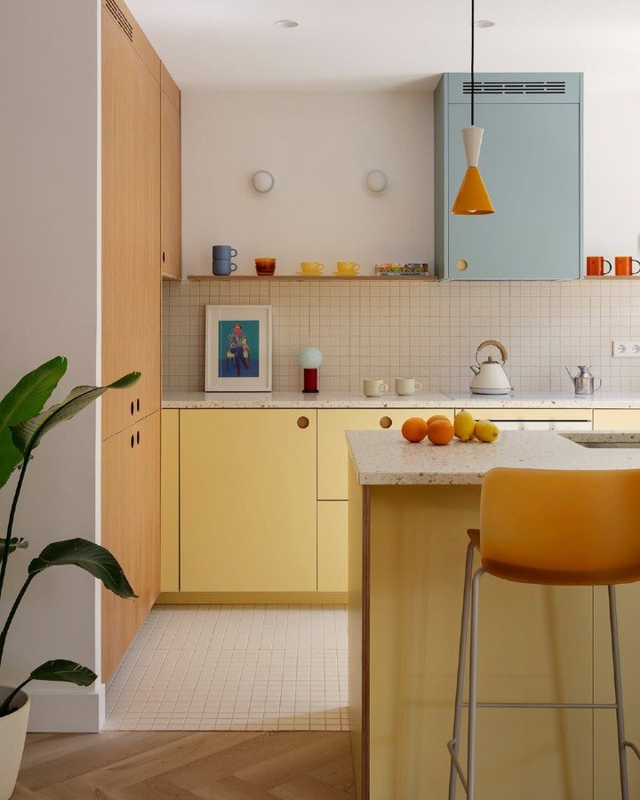 Yellow and blue kitchen cabinets with colorful mugs and citrus fruits on a white countertop.