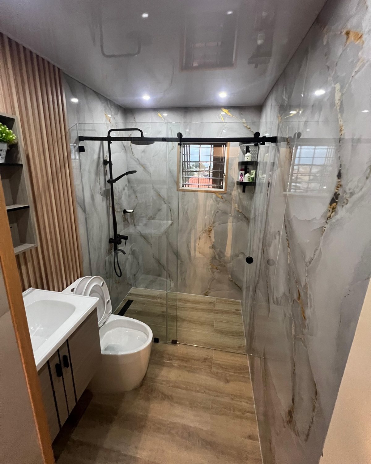 Modern bathroom with marble patterned wall panels, glass shower, and wood-look flooring.
